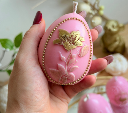 Lumânare decorativă roz în formă de ou cu floare aurie și model floral în relief, ținută în mână, decor de Paște handmade